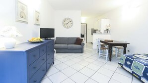 A modern living room with a sofa, a TV, a dining table with chairs, and a clock on the wall. - Il Porto Bilocale 5 (Porto Azzurro)