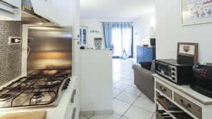 A modern kitchen with a gas stove, a microwave, and a coffee machine. There is a dining area with a blue sofa and a cabinet. - Il Porto Bilocale 5 (Porto Azzurro)