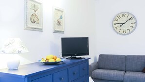 A living room with a blue dresser, a flat-screen TV, a gray sofa, and a wall clock. - Il Porto Bilocale 5 (Porto Azzurro)