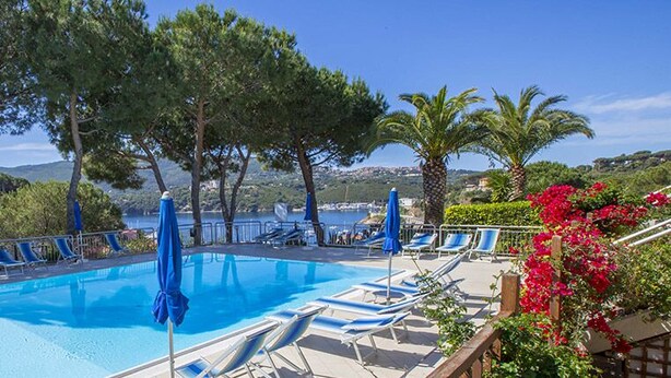 A swimming pool with lounge chairs and umbrellas, surrounded by trees and a view of the sea. - Il Porto Bilocale 13 (Porto Azzurro)