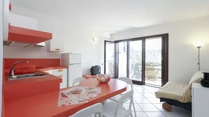 A compact kitchen with a red countertop, a sink, and a refrigerator. There is a dining area with a table and chairs, and a bed with a white headboard. - Il Porto Bilocale 13 (Porto Azzurro)