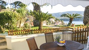 An outdoor patio with a round wooden table, two wooden chairs, and a bowl of fruit. A wooden fence surrounds the patio, and there are palm trees and a view of a lake and buildings in the background. - Il Porto Trilocale 7 (Porto Azzurro)