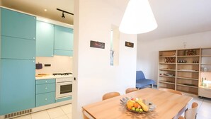 A modern kitchen with teal cabinets, a dining area with a wooden table, and a blue sofa. - Il Porto Trilocale 7 (Porto Azzurro)