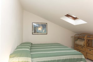 A bedroom with a bed, a framed picture on the wall, a skylight, and a wicker furniture piece.