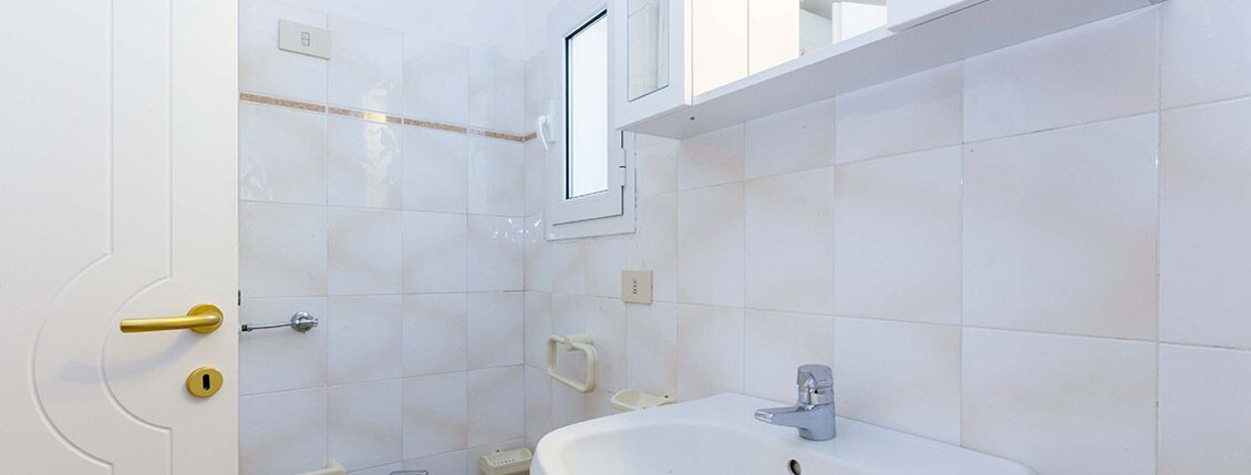 Ein Badezimmer mit weißen Fliesen, einem Waschbecken, einem Spiegel und einem Fenster.