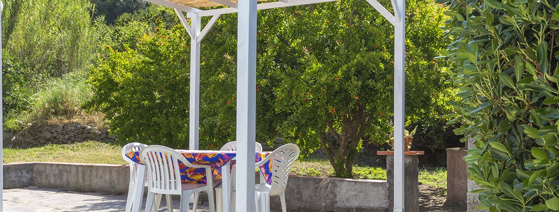 Een overdekt terras met witte stoelen en een tafel, omringd door groen en bomen.