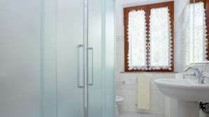 A bathroom with a glass shower door, a toilet, a sink, and lace curtains on the window. - Appartamento Casa Sabrina  (Portoferraio)