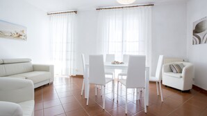 A modern living room with a white sofa, a dining table with chairs, and a large window with sheer curtains. - Appartamento Casa Sabrina  (Portoferraio)