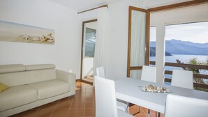 A room with a white sofa, a dining table with chairs, a painting on the wall, and a view of mountains and water through large windows. - Appartamento Casa Sabrina  (Portoferraio)