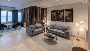 Una sala moderna con dos sofás grises, una mesa de centro y una zona de comedor con una mesa y sillas. Hay dos lámparas de pie y un mapa mundial en la pared.