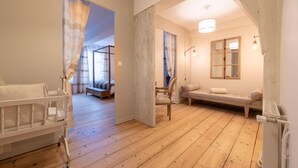 Una habitación con suelo de madera, una cuna blanca, un sofá, una silla y una cama junto a una ventana.