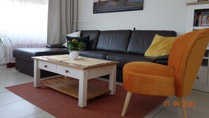 Ein modernes Wohnzimmer mit einer Couch, einem Couchtisch und einem Sessel. An der Wand hängt ein Gemälde und auf dem Tisch steht eine Pflanze.