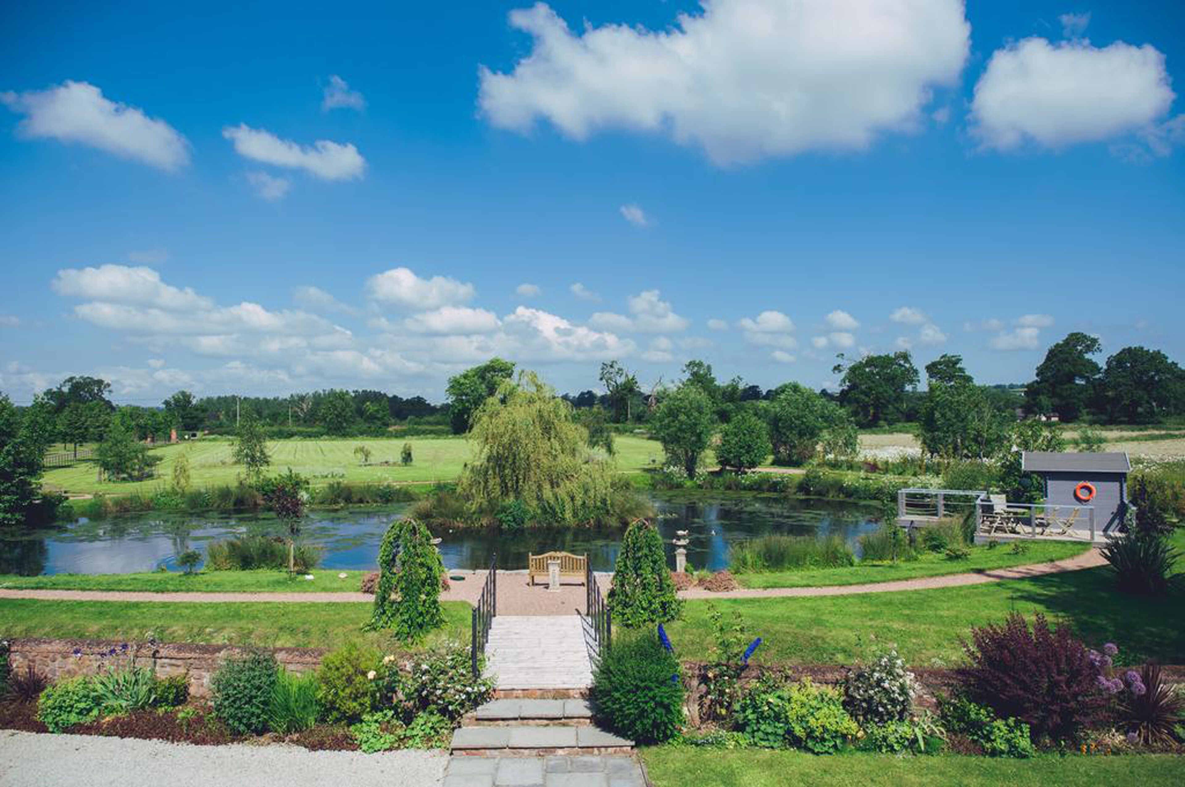 A pathway leading to a pond with a bench and a lifebuoy near a small structure.
