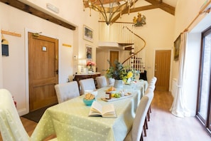 A dining area with a long table set for a meal, a chandelier, and a spiral staircase in the background. - Muddifords Court (Tiverton)