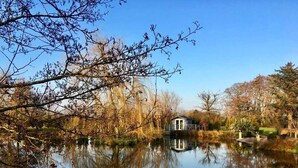 A serene lakeside setting with a small house by the water, surrounded by trees and a clear sky. - Muddifords Court (Tiverton)