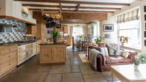 A spacious kitchen with wooden cabinets, a central island, and hanging pots. There is a dining area with a table and chairs, and a cozy seating area with a leather sofa and cushions. - Muddifords Court (Tiverton)