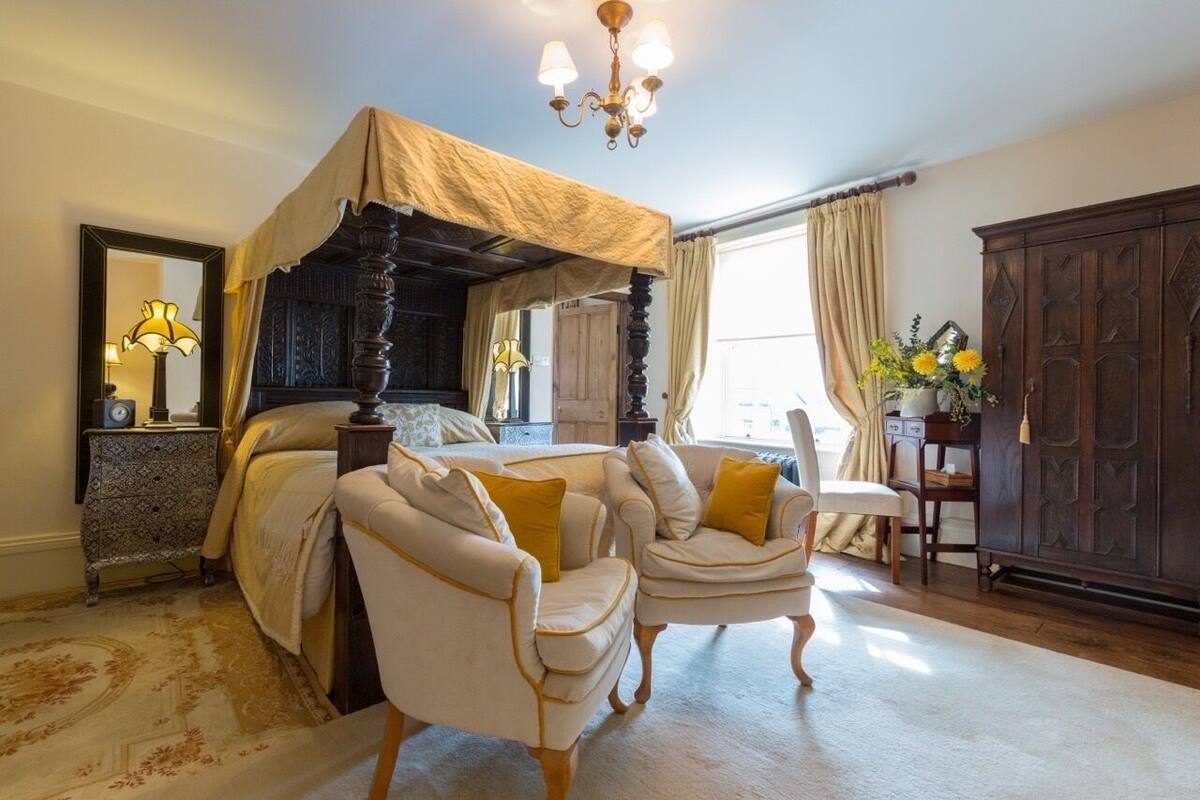 A bedroom with a four-poster bed, two armchairs, a wooden wardrobe, and a large mirror.