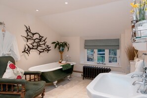 A bathroom with a freestanding bathtub, a large mirror, a green chair, and a window with a view. - Muddifords Court (Tiverton)