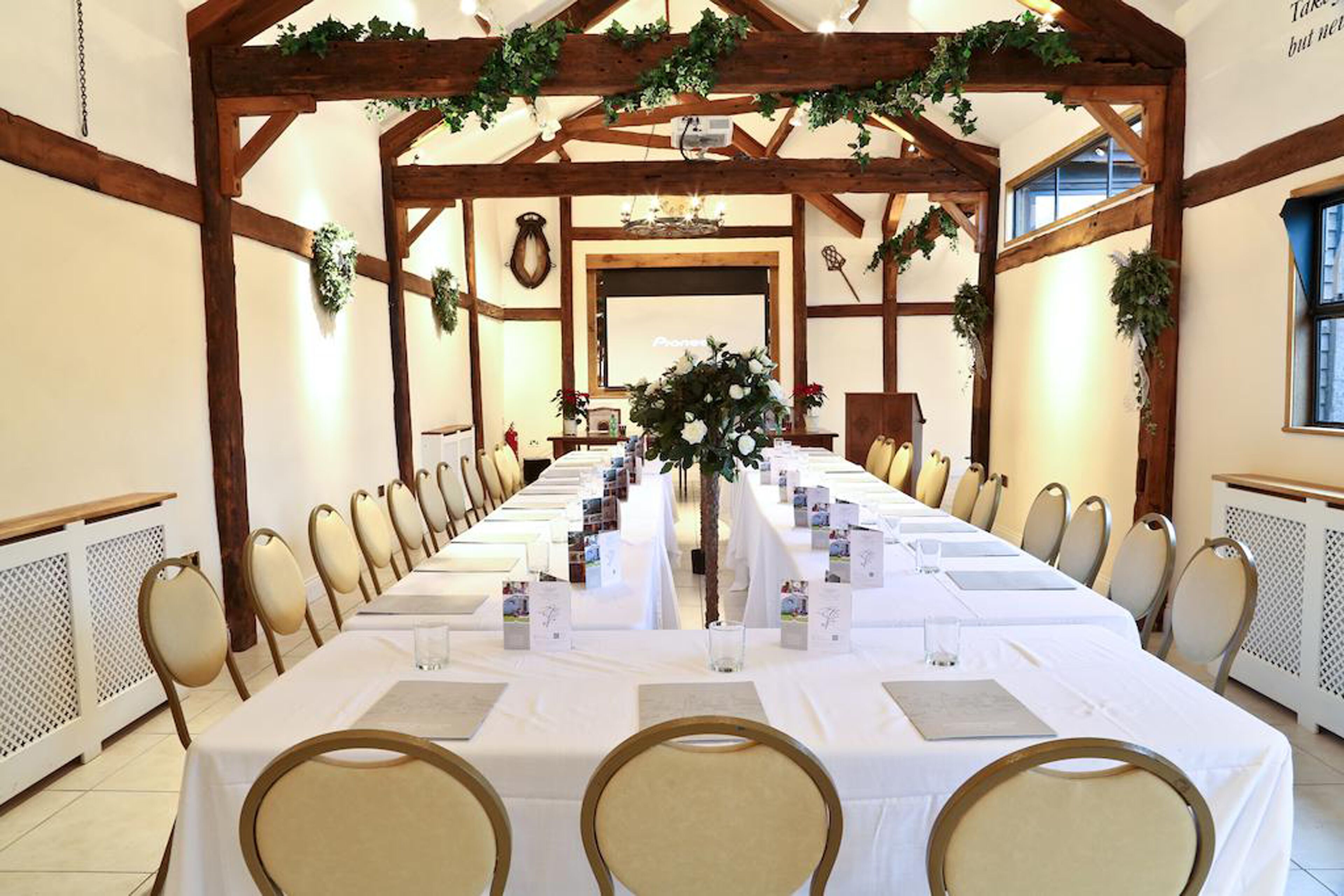 A long dining table set for a formal event, surrounded by chairs, with a wooden ceiling structure and greenery decorations.
