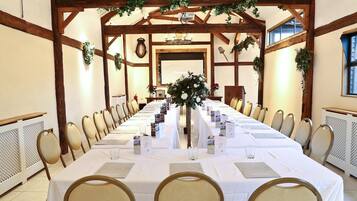 A long dining table set for a formal event, surrounded by chairs, with a wooden ceiling structure and greenery decorations.