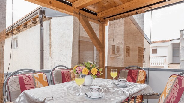 A covered outdoor dining area with a table set for two, chairs with colorful cushions, and a flower vase. - Apartment Banic (Vir)