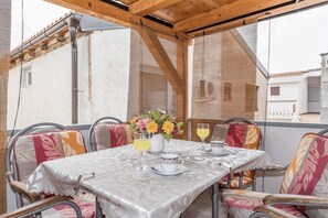 A covered outdoor dining area with a table set for two, chairs with colorful cushions, and a flower vase. - Apartment Banic (Vir)