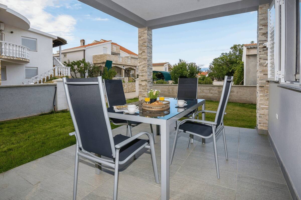 An outdoor patio with a glass table and four chairs, a tray with food and drinks, and a view of neighboring houses.