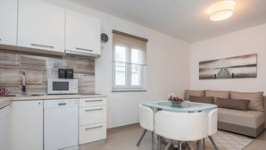 A modern kitchen with white cabinetry, a microwave, and a dishwasher. There's a dining area with a glass table and white chairs. A beige sofa is in the living area, and a painting is on the wall. - Apartments Kosic (Vir)