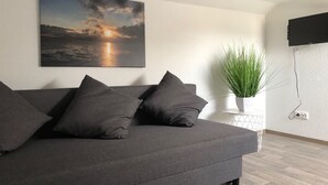 Una sala de estar moderna con un sofá gris, un televisor montado en la pared, una planta en maceta y un cuadro de una puesta de sol.