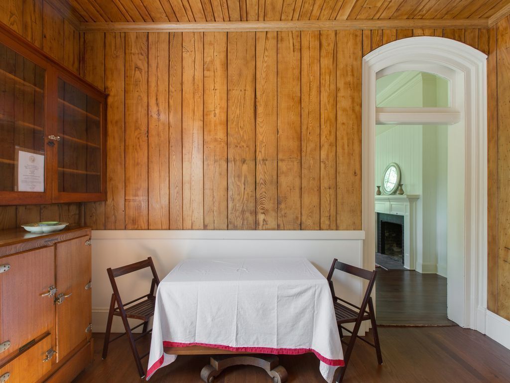 A room with wooden paneled walls, a white table with a red trim, two wooden chairs, a wooden cabinet, and a fireplace visible through an archway.