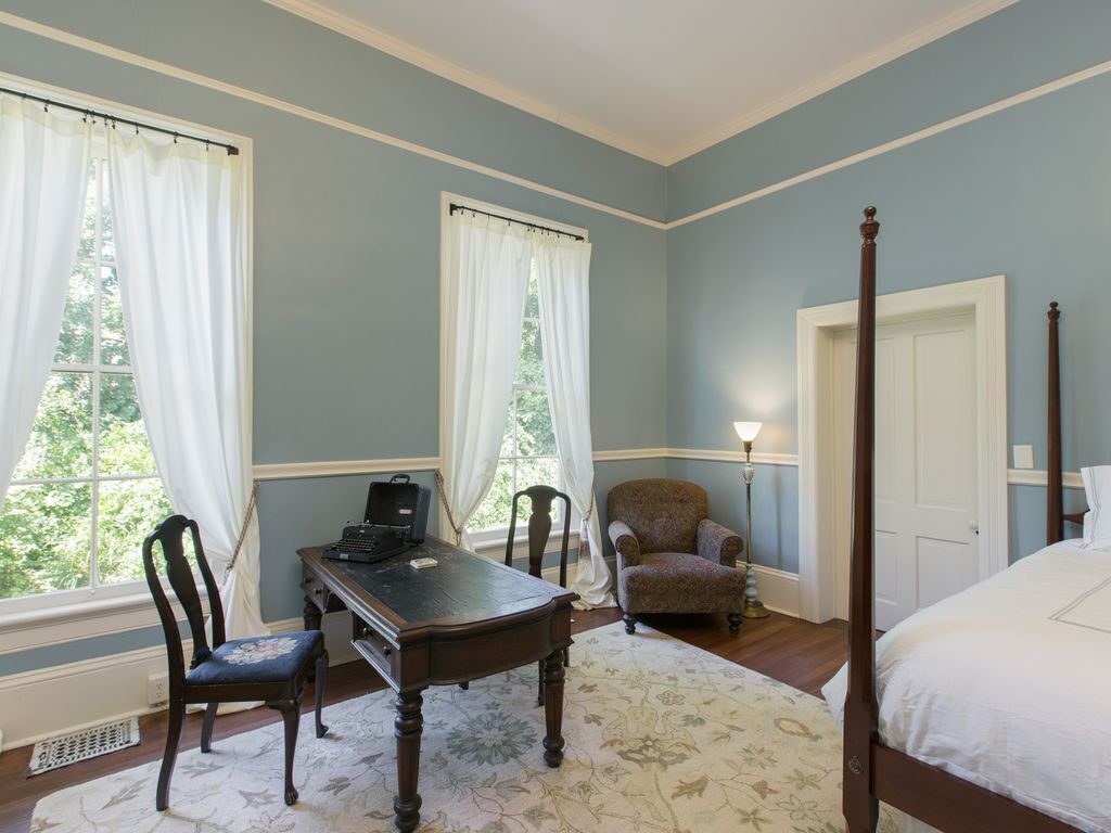 Une chambre à coucher avec un lit à baldaquin, un bureau avec une chaise, une lampe et deux fenêtres avec des rideaux.