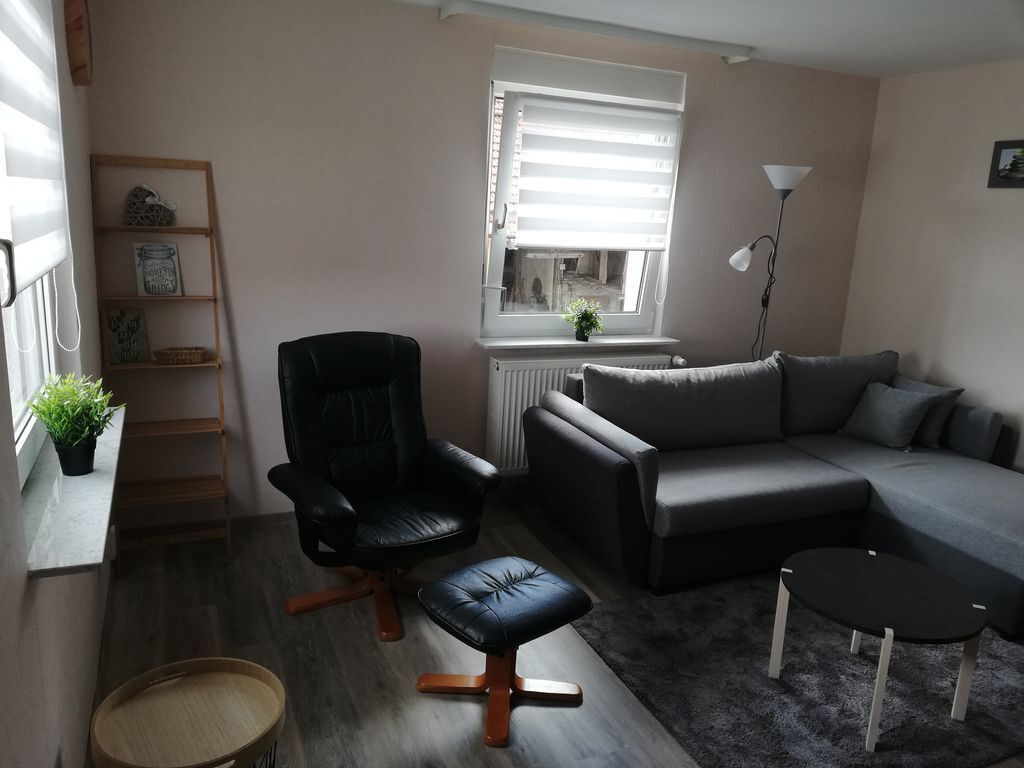 Una sala de estar con un sillón de cuero negro, un sofá gris, una pequeña mesa negra y un radiador cerca de una ventana.