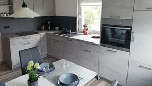 Een moderne keuken met grijze kastjes, een eettafel met stoelen, een hanglamp en een raam met uitzicht op groen.