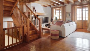 Un escalier en bois menant à une chambre aménagée en mezzanine, un salon avec un canapé et un téléviseur, et de grandes fenêtres dotées de rideaux.