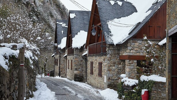 Une rue étroite, recouverte de neige, bordée de bâtiments en pierre dotés de balcons en bois et d’une boîte aux lettres rouge.