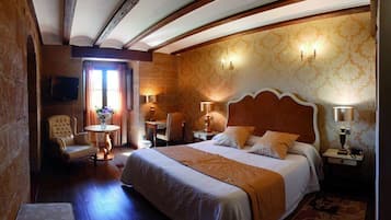 Habitación de hotel con una cama grande, una zona de estar con una silla y una mesa, un televisor montado en la pared y un papel tapiz estampado.
