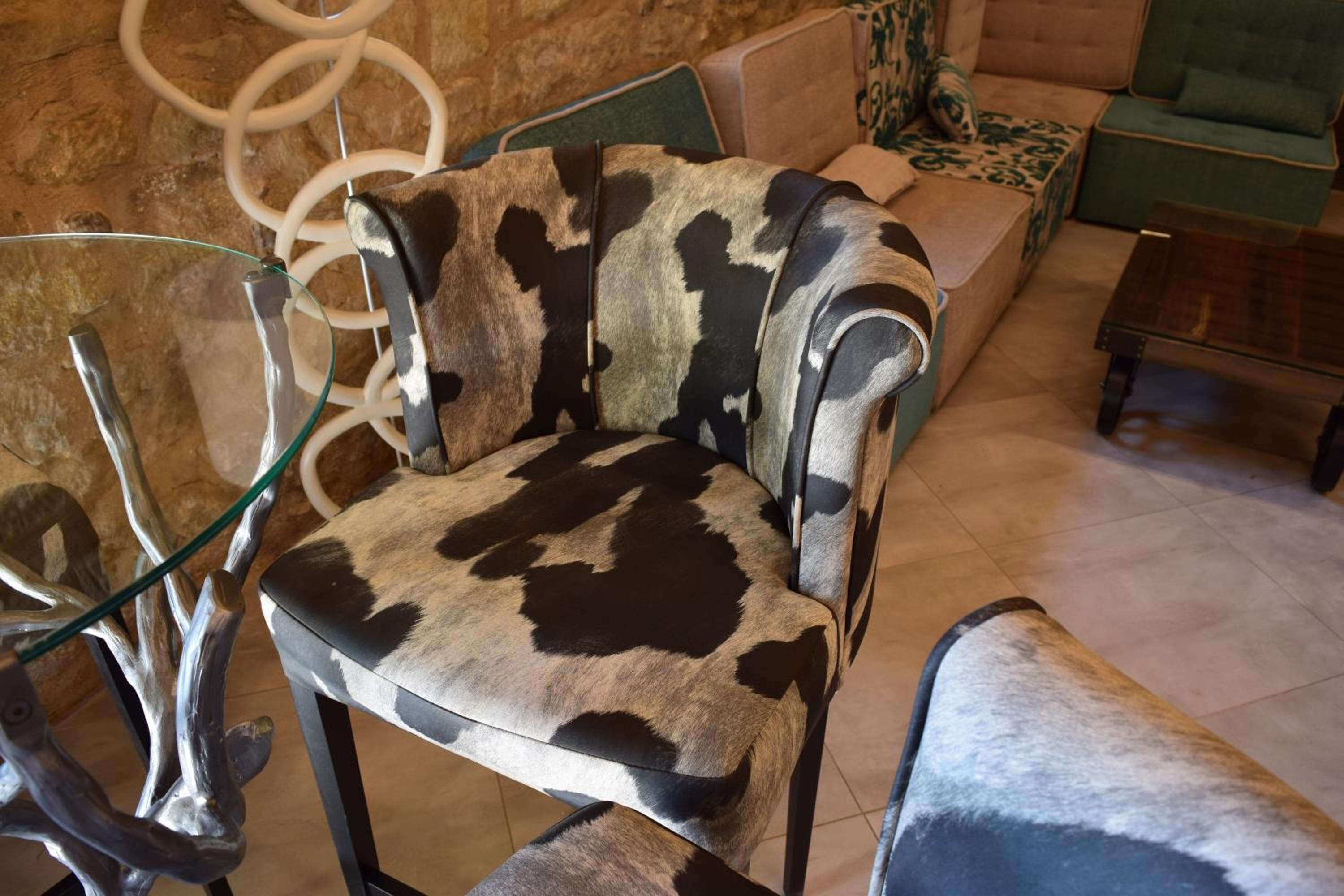 A chair with a cowhide pattern upholstery, a glass table, and a green upholstered sofa in a room with a stone wall.