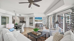 Ein geräumiges Wohnzimmer mit Deckenventilator, einer Kronleuchter und Blick auf den Strand durch große Fenster.