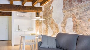 Une cuisine moderne avec des meubles blancs et un four encastré, un canapé gris agrémenté d’un coussin et un mur en pierre au fini rustique.