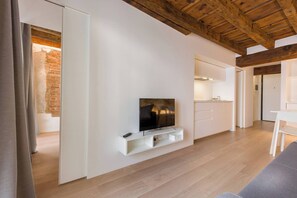 A modern living room with a flat-screen TV mounted on a white wall, wooden flooring, and a visible kitchen area with white cabinets.