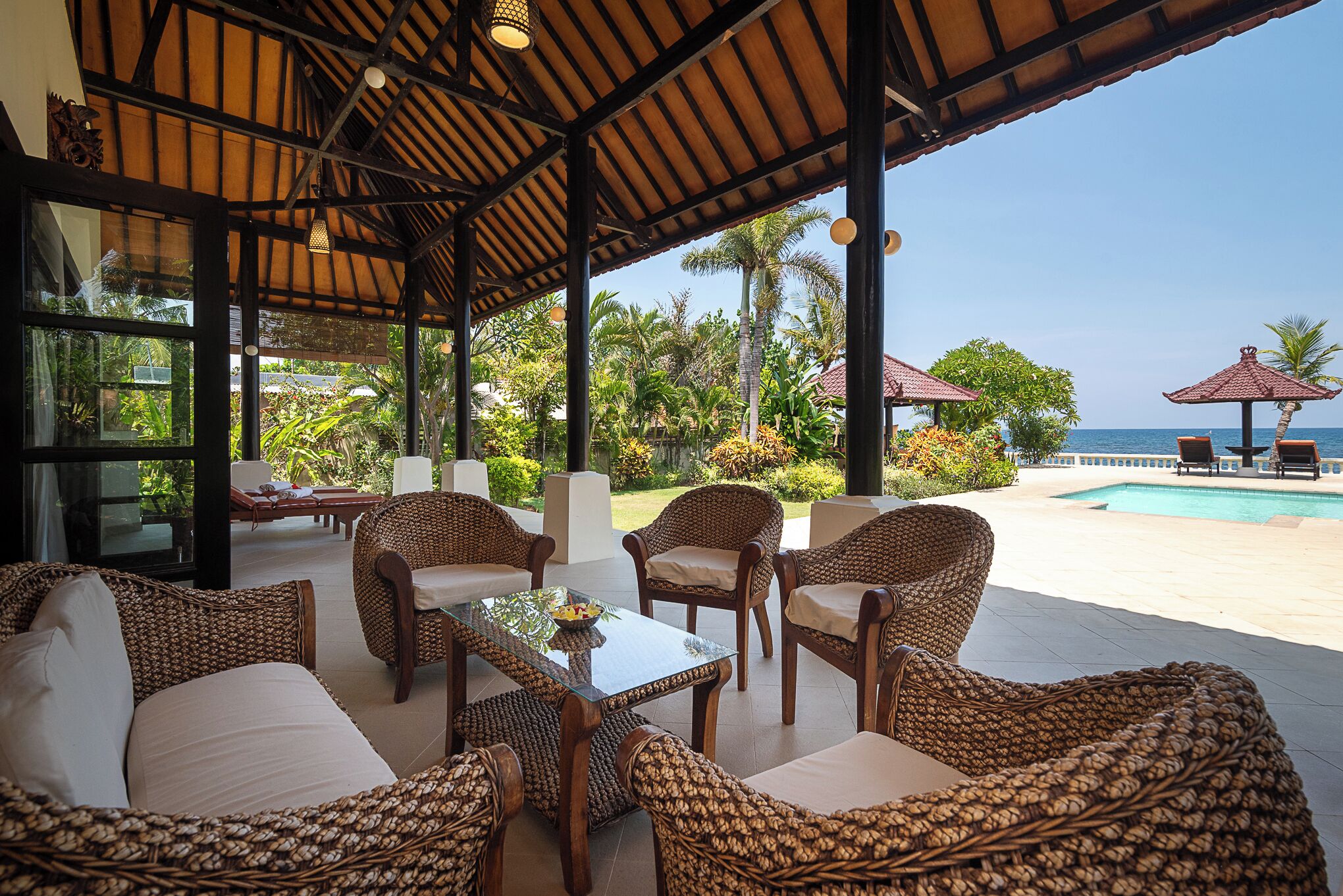 A covered outdoor seating area with wicker furniture, a glass table, and a view of the pool and ocean.