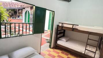 A room with a bed, a bunk bed, a desk, and a view of an outdoor area with a tiled floor and a green door.