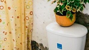 A toilet with a white seat and lid, a yellow floral shower curtain, and a potted plant with yellow flowers on top of a white toilet tank. - Green Door Hostal San Gil - Hostel (San Gil)