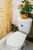 Un inodoro con asiento y tapa blancos, una cortina de ducha amarilla con motivos florales y una planta en maceta con flores amarillas sobre el tanque del inodoro.