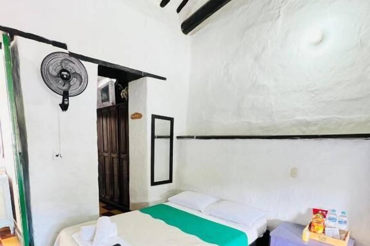 Una habitación de hotel con cama, mesita de noche con lámpara, ventilador y un mueble pequeño.