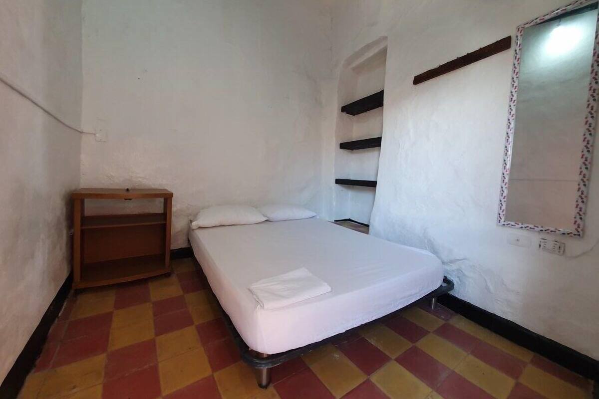 Una habitación pequeña con cama individual, piso a cuadros, una mesita de noche de madera y un espejo en la pared.