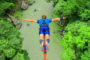 Una persona haciendo bungee jumping con los brazos extendidos sobre un río rodeado de vegetación.