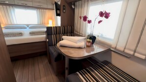 Ein gemütliches Interieur mit einem Bett, einer Sitzecke mit Kissen, einem kleinen Tisch mit Blumenvase und einem Fenster mit Vorhängen.
