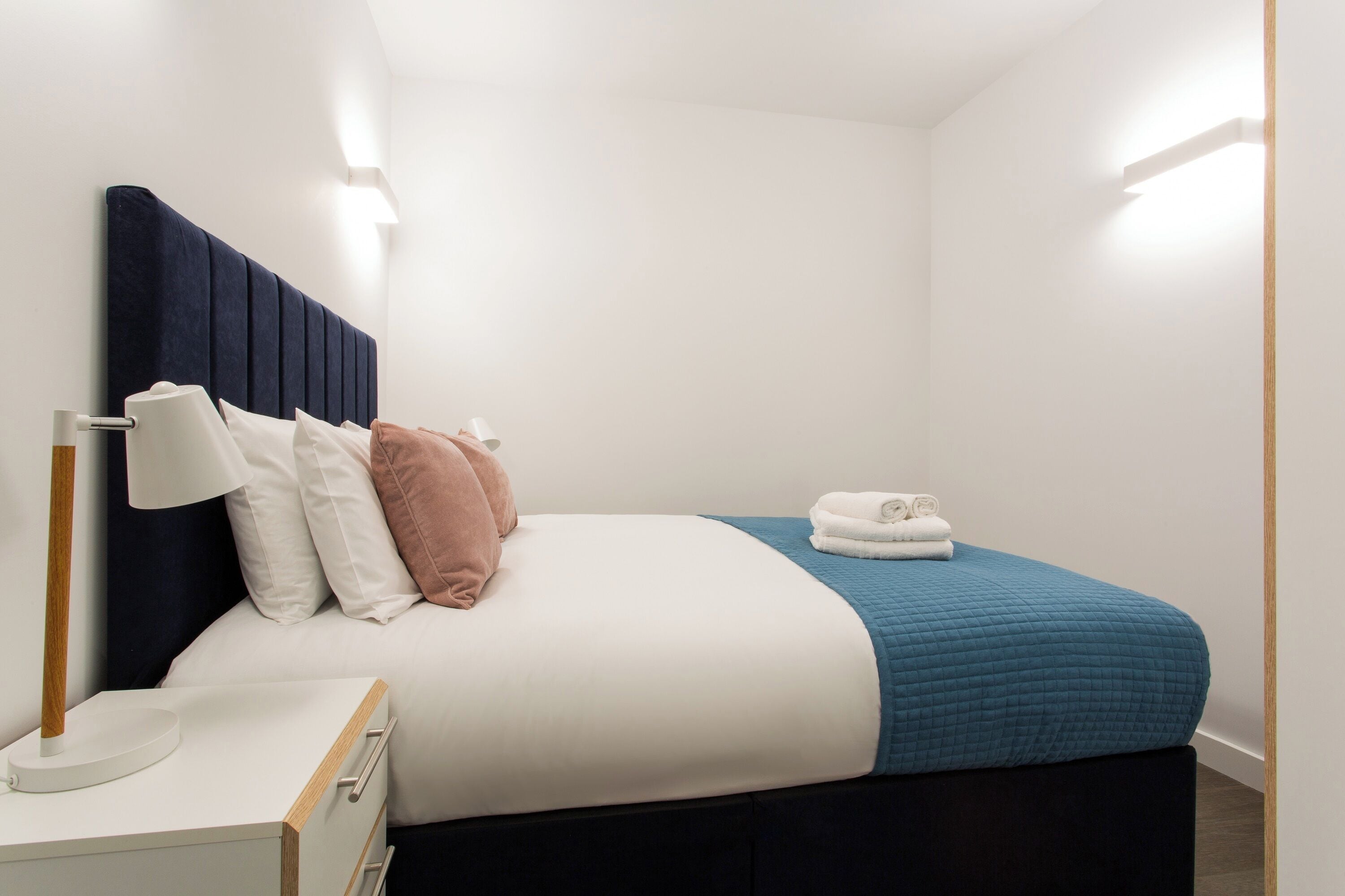 Un lit bien fait, avec une couette bleue, des oreillers blancs et une tête de lit sombre. On y trouve une table de chevet avec une lampe et une serviette pliée.