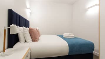 Un lit bien fait, avec une couette bleue, des oreillers blancs et une tête de lit sombre. On y trouve une table de chevet avec une lampe et une serviette pliée.